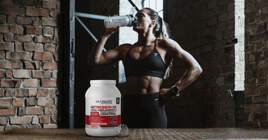 Athletic person in a gym drinking from an Ultimate Nutrition shaker with a large container of Ultimate Nutrition Metressyn RX meal replacement protein powder in the foreground and brick walls and gym equipment creating a fitness focused atmosphere