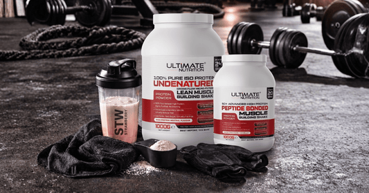 Gym floor scene with two Ultimate Nutrition protein powder containers one ISO Protein neutral flavour and one 50 Advanced High Protein vanilla flavour placed beside a strawberry shaker bottle a scoop of protein powder and black workout gloves with barbell