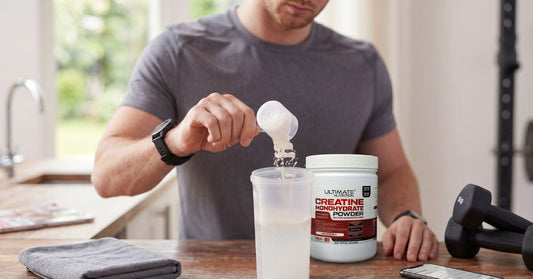 person scooping ultimate nutrition creatine monohydrate powder into a shaker bottle on a kitchen counter with dumbbells nearby to illustrate how creatine works in everyday fitness routines for the article how does creatine work the science explained simpl