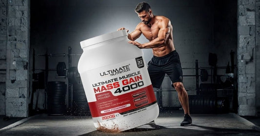 Muscular person in a gym standing behind a large container of Ultimate Nutrition Ultimate Muscle Mass Gain 4000 protein powder with weightlifting equipment and concrete walls in the background creating a strong strength training atmosphere