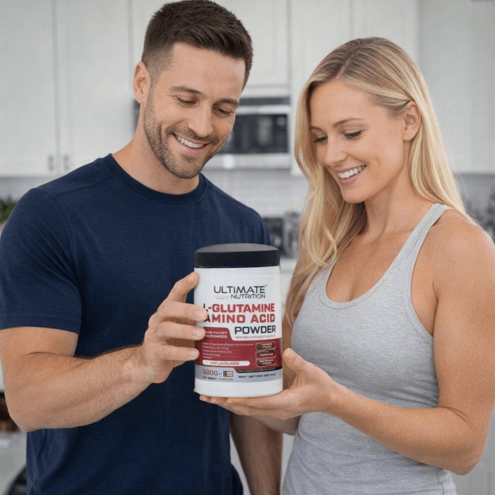 Two people standing in a kitchen holding a container of Ultimate Nutrition L‑Glutamine amino acid powder while discussing post‑workout recovery and wellness
