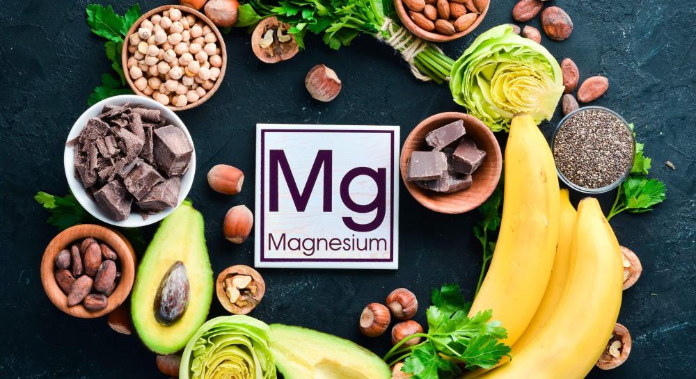 Assorted magnesium‑rich foods including nuts, seeds, leafy greens, bananas, avocado, dark chocolate, and chickpeas arranged around a magnesium sign