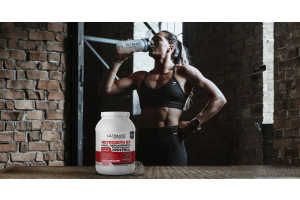 Athletic person in a gym drinking from an Ultimate Nutrition shaker with a large container of Ultimate Nutrition Metressyn RX meal replacement protein powder in the foreground and brick walls and gym equipment creating a fitness focused atmosphere