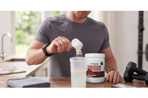person scooping ultimate nutrition creatine monohydrate powder into a shaker bottle on a kitchen counter with dumbbells nearby to illustrate how creatine works in everyday fitness routines for the article how does creatine work the science explained simpl