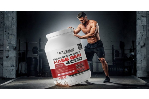 Muscular person in a gym standing behind a large container of Ultimate Nutrition Ultimate Muscle Mass Gain 4000 protein powder with weightlifting equipment and concrete walls in the background creating a strong strength training atmosphere